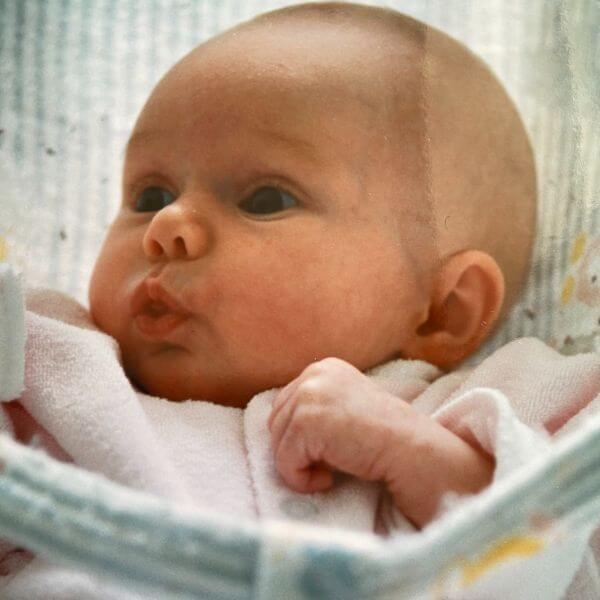 Newborn Laura lying in a baby seat. She is wearing a pale pink onesie and pursing her lips, making a cute pout.