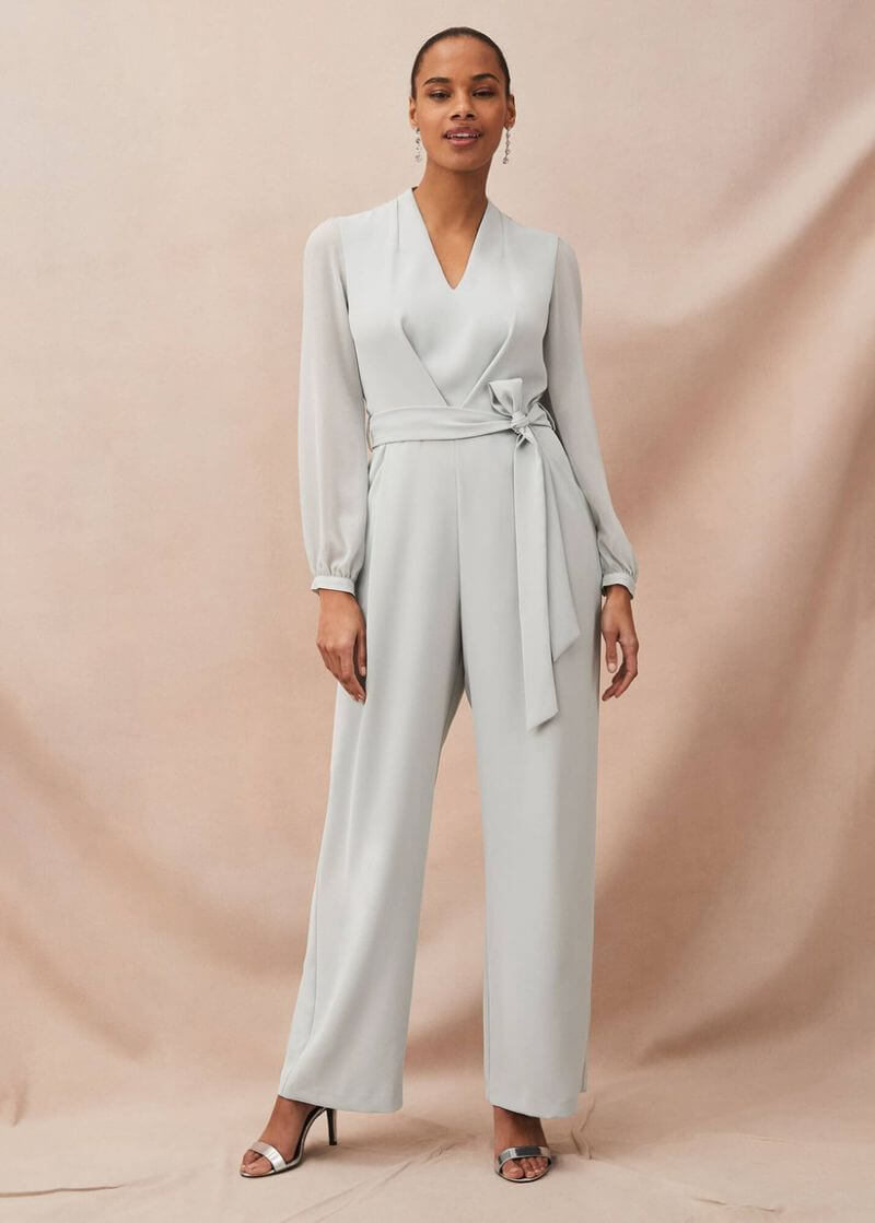 Young, black woman wearing a pale blue jumpsuit with flared legs and long sleeves. The jumpsuit is tied at the waist. Her dark hair is styled back and she is smiling at the camera. She also wears long, dangly earrings.