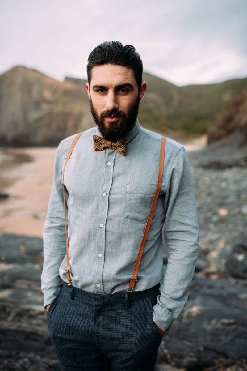 White man with a dark brown beard and hair wearing a blue shirt and dark trousers. Light brown braces hold up his trousers and he is wearing a bow tie to match. His hands are in his pockets.