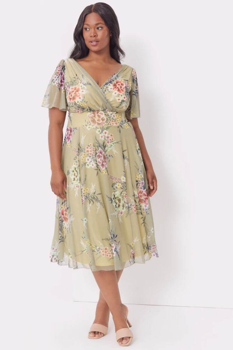 Young black woman with dark hair wearing a green, floral, knee-length dress. The material is floaty and pretty. She is wearing nude high-heeled sandals.
