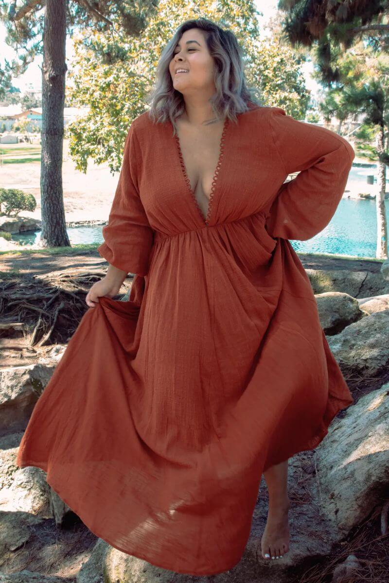 Plus-size white woman with short lilac hair. She is standing on some rocks in front of a pool of water and wearing a floor-length, bohemian-style red dress that plunges at the cleavage. The material looks light and summery. She is smiling and holding her skirt on one side. Her other hand is on her hip.