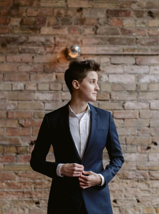 Androgynous white person with short, tussled brown hair wearing a white shirt and navy blue suit jacket, stood in front of a brick wall. They are buttoning their suit jacket.