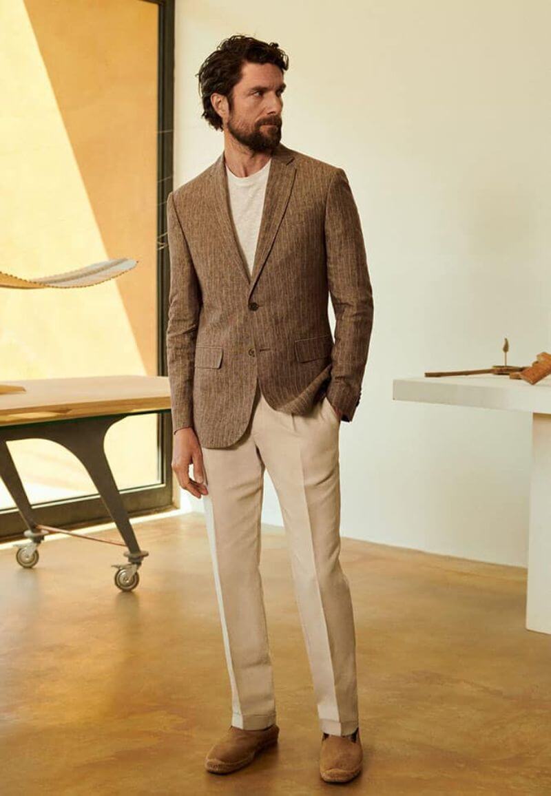White man with brown beard and slightly longer, shaggy hair wearing beige trousers and a white t-shirt, covered by a brown pin-stripe suit jacket. He wears brown suede shoes and one of his hands is in his pocket.