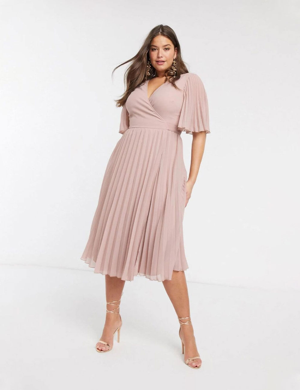 White woman with long brown, wavy hair standing in a pale pink pleated dress with butterfly sleeves. She wears pale pink high-heeled sandals to match.
