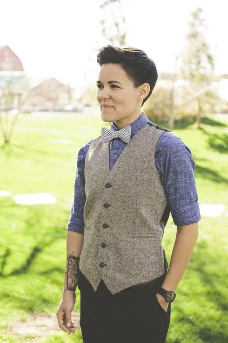 Young, androgynous person with short brown hair, stood smiling and looking to the side. They are wearing dark trousers, a blue shirt with the sleeves rolled up to the elbows, a beige waistcoat and a white bow tie. One hand is in their pocket, and on the other hand they have a tattoo on their wrist.
