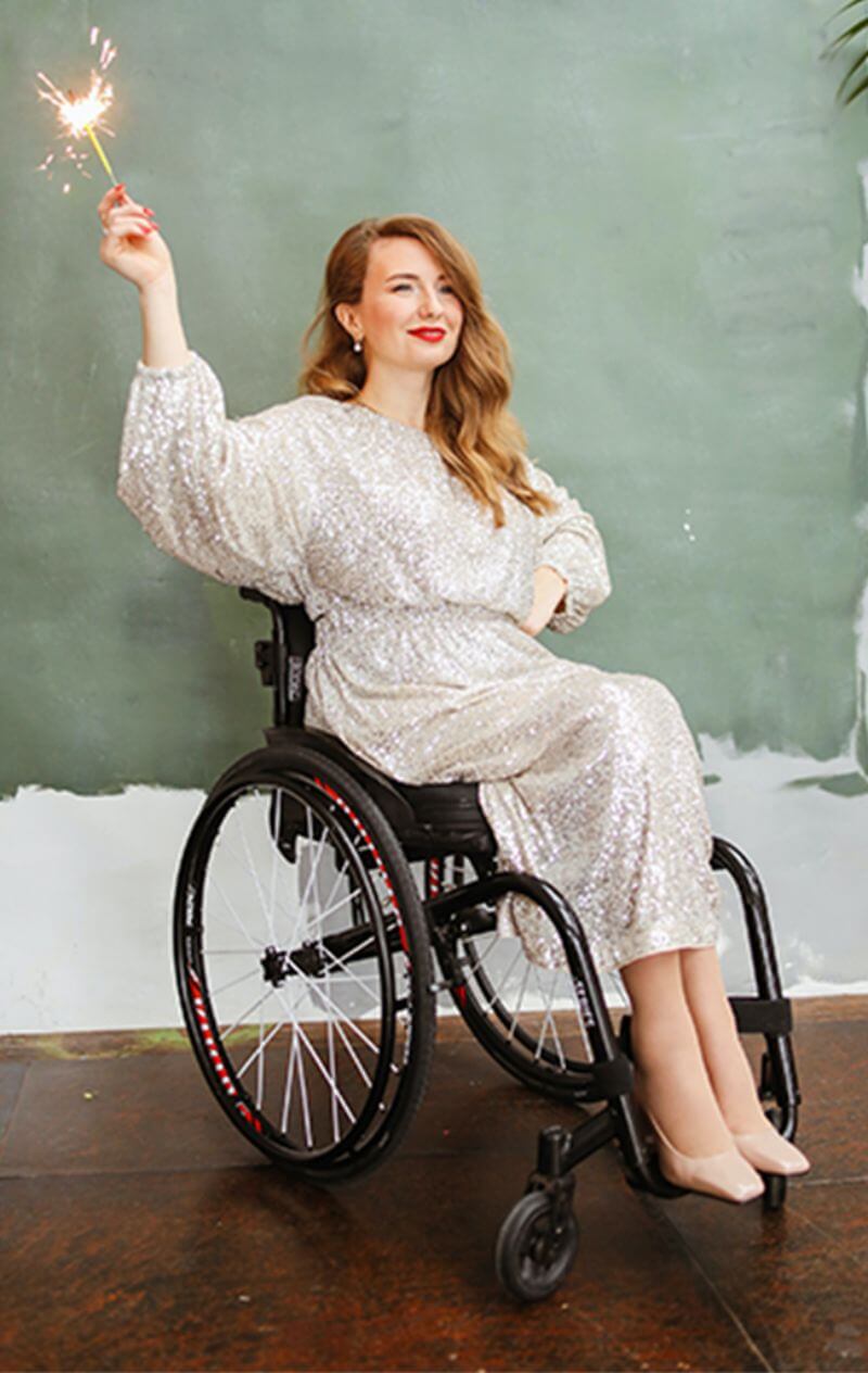 White woman with long, wavy auburn hair sits in her wheelchair. Her dress is silver and covered in sparkles and is accompanied by nude closed-toe shoes. Her lips and nails are painted red and she is smiling, holding a sparkler up with her right hand.