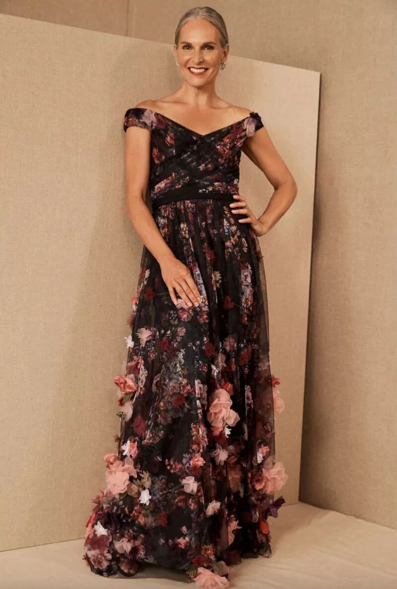 Older white woman with silver, pinned-back hair. She is smiling widely and wearing a long, off-the-shoulder gown that is predominantly black but covered in red and pink textured flowers. The flowers get bigger the lower on the dress they are placed. Her hand is on her hip.