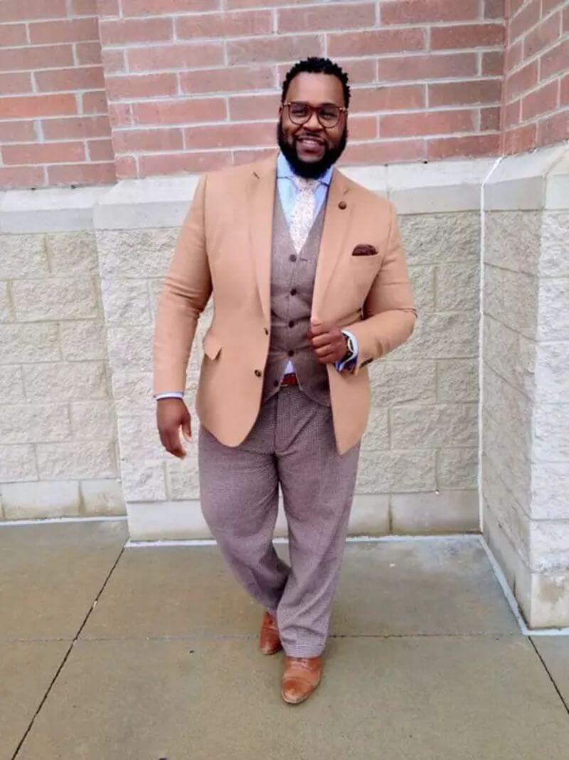 Plus-size black man with black hair and beard in glasses, grinning widely. He wears matching brown trousers and waistcoat with a lighter brown jacket. His shirt is pale blue and his tie is baby pink. He wears brown leather shoes.