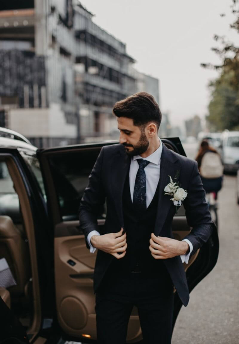 White man with dark hair and beard stepping out of a car. He is wearing a black suit and waistcoat, white shirt, black tie and a white rose buttonhole. He is in the process of adjusting his jacket.